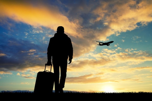 A silhouette of a person pulling a suitcase on wheels in the foreground of a sunrise, with the silhouette of a jet in flight in the sky in the background.